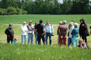 The image shows a group of people gathered outdoors, listening attentively to a conversation or presentation.