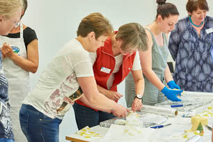 The image depicts several individuals collaborating on a project at a table, likely within the realm of crafts or art.