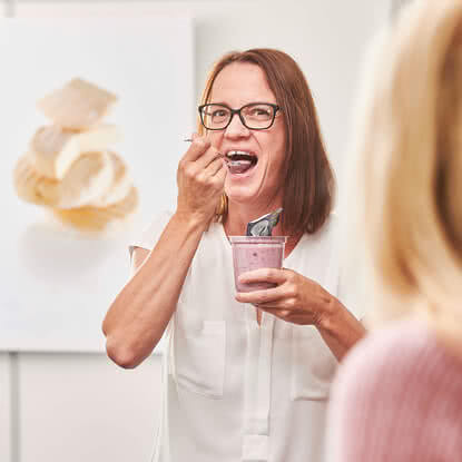 The image features a woman smiling as she enjoys a purple dish from a cup, while looking directly at the camera.