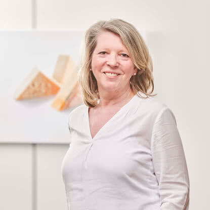 The image features a woman in a white top smiling, with a work of art showcasing pieces of our artisanal Allgäu cheese in the background.