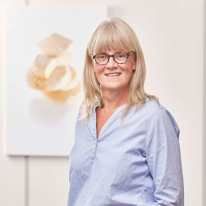 The image features a smiling woman with blonde hair and glasses, standing in front of a blurred piece of art.