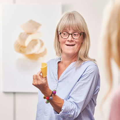 The image features a smiling woman wearing glasses, holding a piece of artisanal organic cheese from Allgäu in her hand while chatting with a customer.