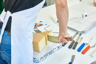 The image depicts a artisan expertly slicing a wheel of organic Allgäu cheese with a large, sharp knife, surrounded by a selection of professional cheese-cutting tools.