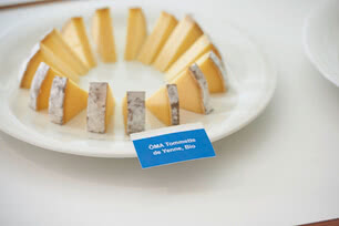The image presents an assortment of wedges on a serving plate, each accompanied by a label reading ‘ÖMA Tommette de Yenne, Organic’.