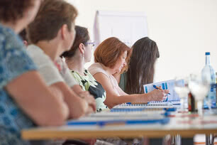 The image depicts several women sitting at a table during a meeting or training session, with one of them taking notes.