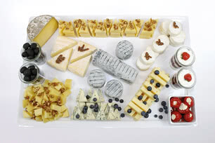 The image presents a diverse spread of artisanal cheeses, cured olives, fresh seasonal fruit and house-made desserts, all thoughtfully arranged on a single board.