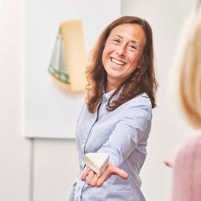 The image depicts a smiling woman offering a piece of our artisanal Allgäu organic cheese to a friend.