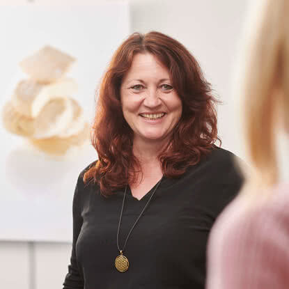 The image features a smiling woman with red hair posing in a brightly lit room. Blurred objects are visible in the background.