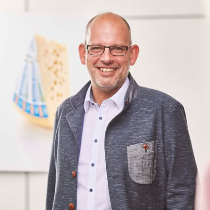 The image features a man smiling while wearing a grey blazer; in the background, a wheel of our organic Allgäuer Bergkäse can be seen.