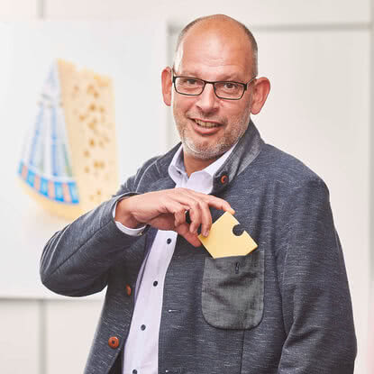 The image features a smiling man cradling a wedge of our handcrafted organic cheese in his jacket pocket, with our aging room visible in the background.