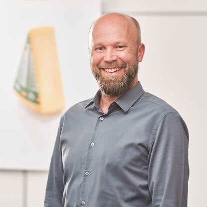 The image features a smiling, bearded man wearing a grey shirt and standing before a light background alongside a piece of our artisanal organic cheese.