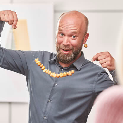 The image features a man smiling while holding up a necklace crafted from wooden beads.