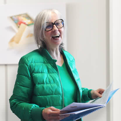 The image presents a smiling woman with grey hair and glasses, wearing a green outfit and holding a booklet in her hand.