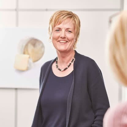 The image features a smiling woman standing in a bright room, with artisanal Allgäu cheese displayed in the background.