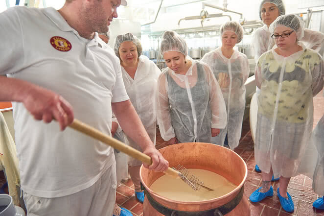 The image shows a group of enthusiasts observing a master cheesemaker at work in our Allgäu cheese-making workshop.
