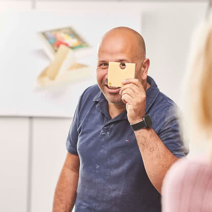 The image features a smiling man holding a piece of our artisanal Allgäu cheese in front of his face.