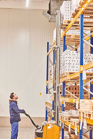 The image depicts a warehouse associate operating an electric hand pallet truck and scanning the tall shelves lined with crates.