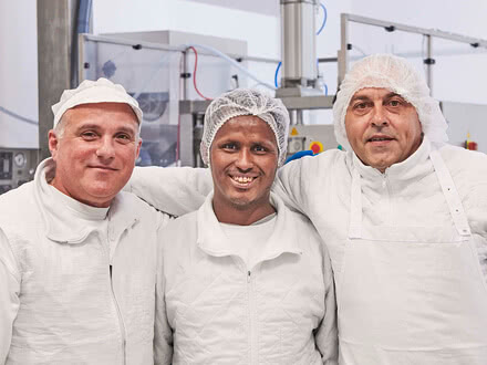 Behind the scenes at our Allgäu creamery, three of our cheesemakers—dressed in white overalls and hairnets—share a moment of pride in our spotless production facility.
