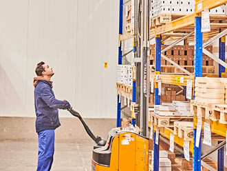 The image depicts a warehouse associate operating an electric forklift and glancing upward, likely to retrieve a pallet of freshly delivered, certified organic ingredients.