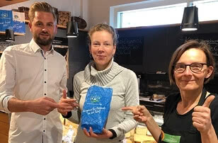 The image shows three individuals standing around a blue parcel, smiling as they point at it.
