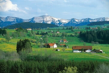 Our Allgäu region is captured in this image: an idyllic rural landscape with green meadows, scattered farmhouses and snow-capped mountains in the background.