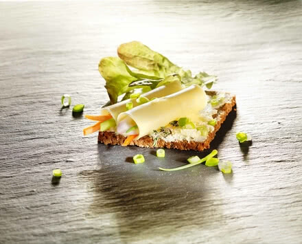 The image presents a sandwich topped with our artisan Allgäuer Bergkäse, fresh organic vegetables and a hint of aromatic herbs.