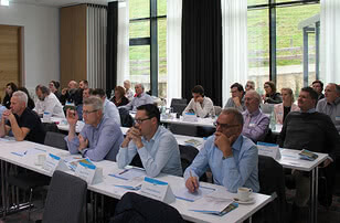 The image shows participants in a seminar or conference listening attentively and taking notes.