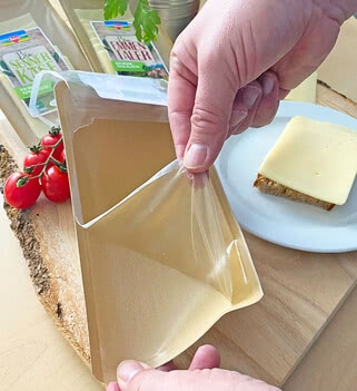 The image shows a customer opening a packet that probably contains our handcrafted Allgäu organic cheese, while beside it lies a slice of bread topped with the same cheese and accompanied by fresh, ripe tomatoes.