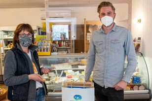 The image shows a woman and a man in a shop; the woman is holding a food item and the man is holding a bag of shopping. Both are wearing masks.