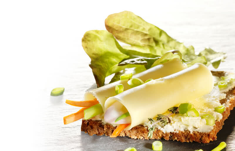 The image presents a sandwich topped with our artisan Allgäu cheese, a selection of crisp organic vegetables, and freshly chopped herbs.