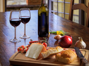 The image presents two glasses of red wine, a bottle of wine, and an assortment of food items such as cheese, bread, tomatoes, and onions on a table.
