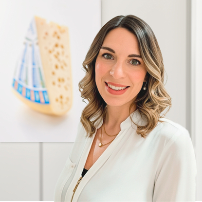 The image features a smiling person standing next to a plate of our artisanal Allgäuer Bergkäse, a raw-milk semi-hard cheese aged in traditional cellars.