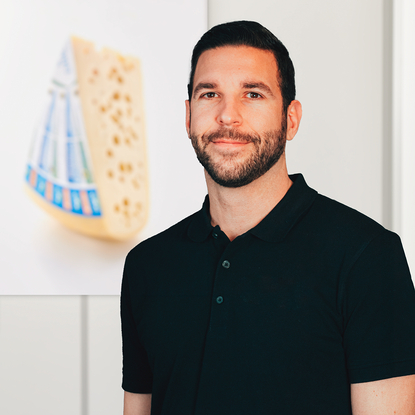 The image features a man in a black polo shirt standing before a backdrop of artisanal cheese, evoking the time-honored dairy craftsmanship of Allgäu.